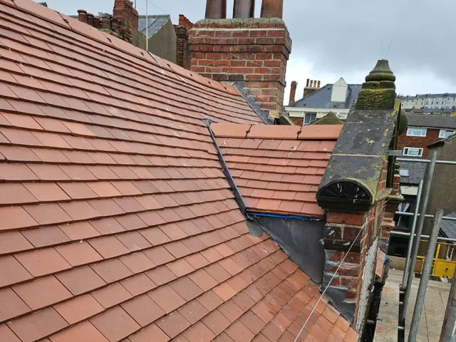 Red clay tiled roof with a lead-lined valley running down beside an old chimney stack, with scaffolding and terraced houses behind